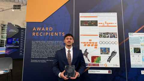 Haukapuanui Vercoe holds his award in front of posters at the 2025 Esri User Conference in San Diego, US
