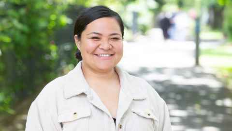 Morgan Patii-Kauhiva wears beige jacket and smiles to camera while standing in University gardens on a sunny day.
