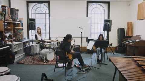 Three students sitting in a circle. One person is playing drums, while other two people play guitar.