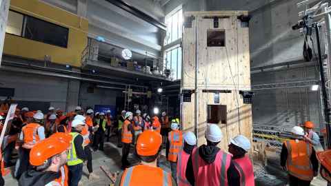 People in high-vis jackets and hardhats inside a large concrete testing hall looking at a two storey structure made from cross-laminated timber