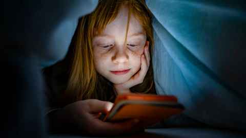 A young child is looking at a glowing mobile phone screen under a sheet in the dark.