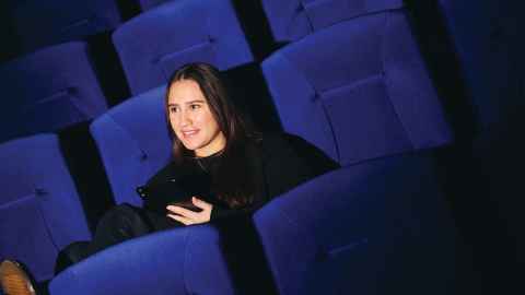 Woman in dark clothing sitting in a blue fabric lecture hall seat, holding a tablet in her hand.
