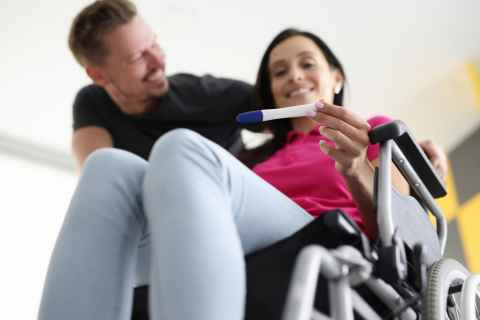 Woman in wheelchair holding pregnancy test and male partner behind her smiling