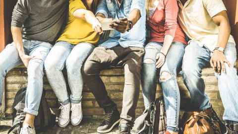 A group of five young people seated outside looking at a phone held by the person in the middle.