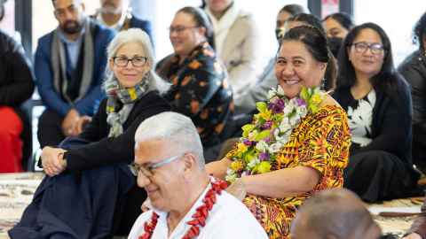 Everyone is seated, and the photo is taken at an angle, rather than directly in front of people. Dawn wears a striped scarf and dark clothing, her arms around her knees. She is smiling slightly, head turned towards Jemaima who sits next to her. Jemaima’s head is turned also, grinning at someone we cannot see. She is wearing a brightly coloured Pacific print dress, and an ula made of various flowers, her hands with traditional tattoos on her knees. Siaosi Gavet sits one level down in front of her, wearing a white shirt and an ulafala. He faces ahead, head tilted down as he smiles. Others sit behind them in the background.