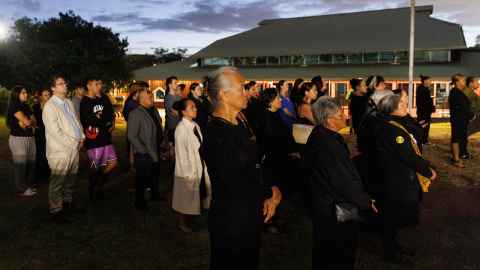 Three kuia stand in the foreground facing to the right at the dawn ceremony. The roof of the whare kai of Waipapa marae is visible in the background, those gathered standing between the kuia and the whare kai.  