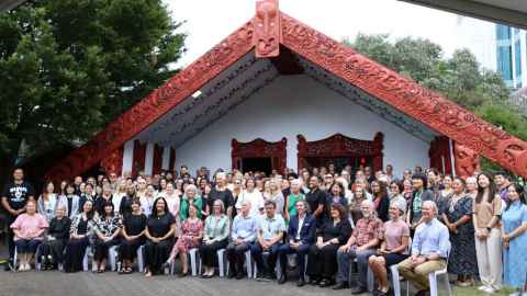 Staff of Education and Social Work and others in front of the Waipapa Marae wharenui. Front row are seated, and include Pro Vice-Chancellor Māori Te Kawehau Hoskins, Deputy Vice-Chancellor Operations Adrienne Cleland, Provost Valerie Linton, Dean of EDSW Mark Barrow. The seven or eight rows behind are standing.