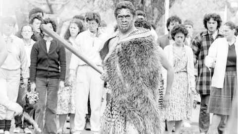 Students protesting outside the building housing University leadership. Foreground a male student in full tā moko with a staff, wearing korowai and piupiu. Middleground rows of students singing, female students in front, some holding flowers. Trees lining Symonds Street in the background.