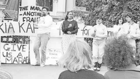 Five students singing in protest outside the building housing University leadership, flanked by protest signs: The Marae - another fraud; Give us a Marae; Kia kaha (balance of banner obstructed by bodies).