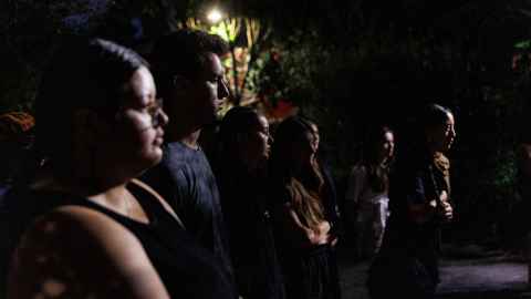 Mostly in the dark, a receeding row of tauira Māori stand in profile, looking beyond the photo bounds to the viewer’s right, with just their faces and hands reflecting the light ahead of them. In the background, the branches of a tree glow from a streetlight.