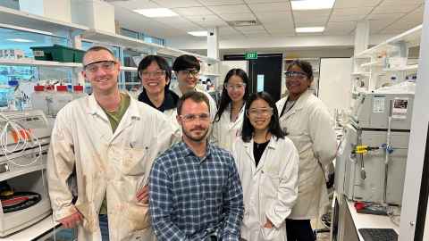 Alan Cameron seated in the lab in plaid shirt, surrounded by his team including students