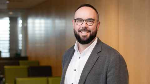 A head shot of Deputy Dean Andrew Patterson. He has dark hair and a beard and wears glasses. He is wearing a dark grey jacket and white shirt.