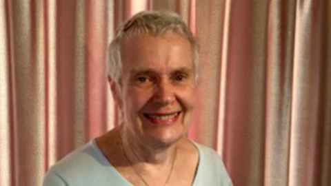 A short-haired woman wearing a white top and gold necklace, standing in front of a pink curtain.