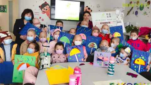 A group of small children wearing masks and holding up drawings, with two adults in the background.