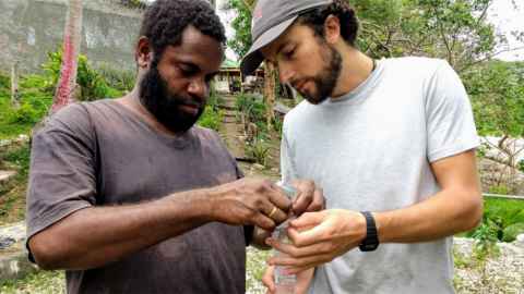 Two men in t-shirts outside, with trees in the background, working on something we cannot see.