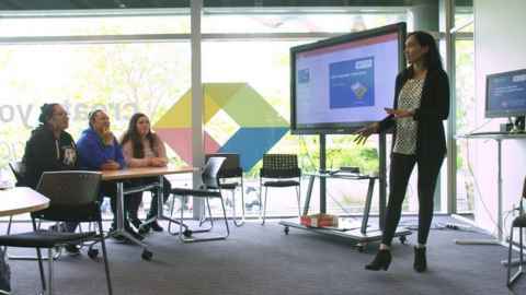 A woman at the front of a classroom, beside a screen, talking to a small group.