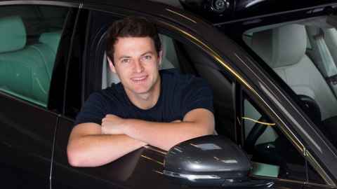 A dark-haired young man leaning out of the driver's side window of a car.