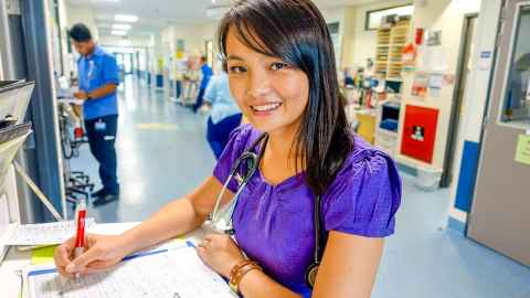 A young woman with a stethoscope around her neck, doing paperwork in a hospital corridor.