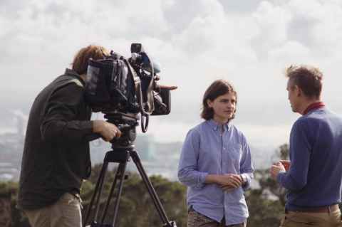 A young woman in a blue shirt talking to an older man. Another man is behind a professional-looking video camera, filming them.