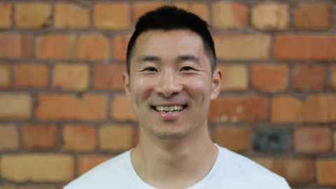 A smiling man in a white t-shirt, in front of a brick wall.
