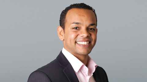 A smiling young man wearing a pale pink shirt and dark jacket.
