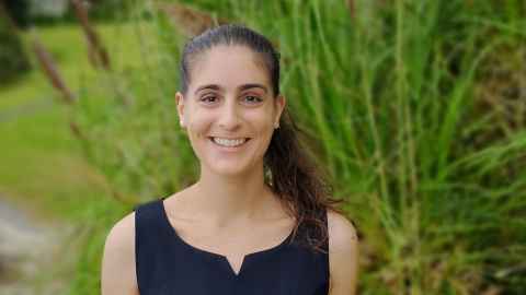 A dark-haired woman, smiling, with trees and bushes in the background.