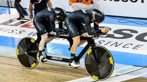 Two women on a tandem bike, wearing black outfits with "New Zealand" on them.
