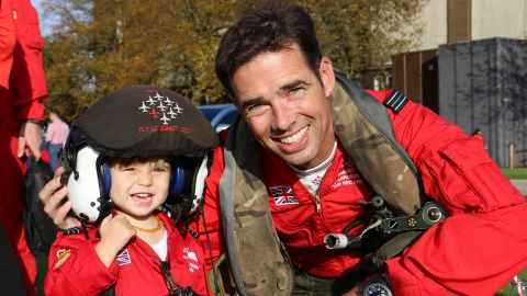 A smiling man in red flight gear, with a small child.