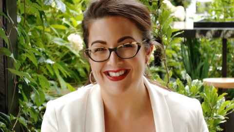 A brown-haired woman wearing a white jacket and red lipstick, with greenery in the background.