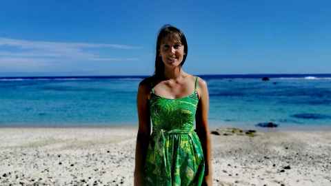A dark-haired woman wearing a green and yellow sundress, standing on a beach.