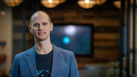 A man in a black t-shirt and blue-grey suit jacket, with a TV screen on a wall behind him.