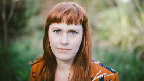 A woman with red hair and blue eyes, with greenery in the background.