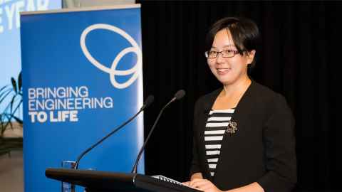 A young woman in a striped top and black jacket at a microphone, next to a sign that reads, "Bringing engineering to life".