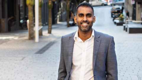 A smiling man in a white shirt and grey suit jacket, standing in a city street.