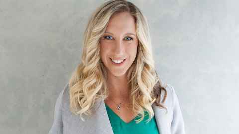 A woman with curly blonde hair wearing a green top and grey jacket.