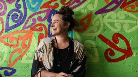 A smiling, dark-haired woman standing in front of a bright green wall with koru and other designs painted on it.