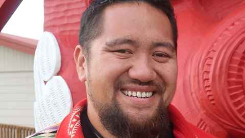A man with a beard, standing in front of Māori carvings.