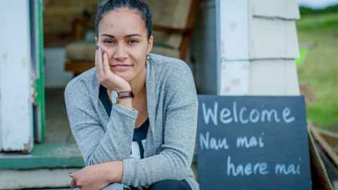A woman in a grey cardigan sitting on a step next to a sign that reads "Welcome, nau mai, haere mai".