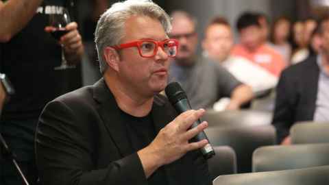 A man with red-rimmed glasses, holding a microphone, sitting among an audience.