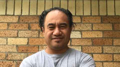 A man in a grey t-shirt standing in front of a brick wall.