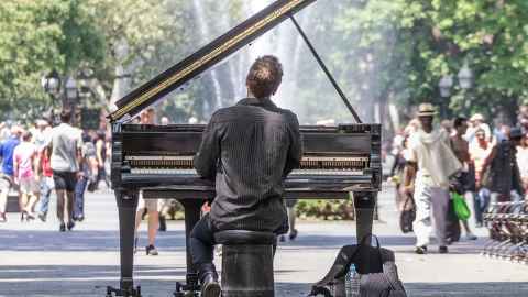 A man playing a grand piano in a busy area with a fountain and trees.
