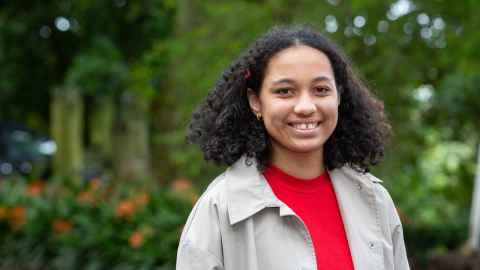 Amalie stands in a university garden and smiles to camera. She wears a red top and beige jacket.