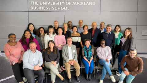 Group of people sitting and standing in front of a wall that has 'the University of Auckland Business School' written on it.