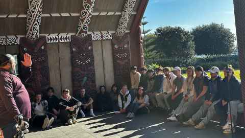 A group of students listen to a Māori speaker inside a marae.
