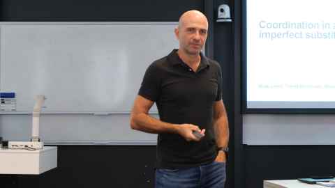 A man at the front of a classroom wearing a black polo t-shirt and jeans 