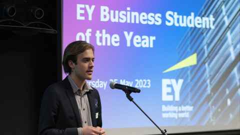 A man standing at a podium talking into a microphone. Behind him is a screen that says 'EY Business Student of the Year'.