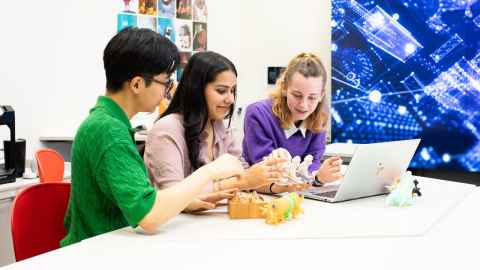 Group of three students sitting at a table which has a laptop on it and colourful objects.