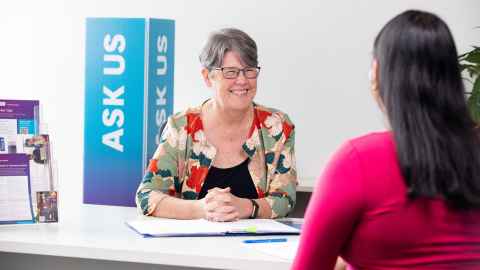 Careers Advisor sits in front of an 'Ask Us' sign, happily conversing with a student.