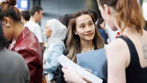 Two people in a busy indoor setting, holding documents and engaged in conversation, with others visible in the background.