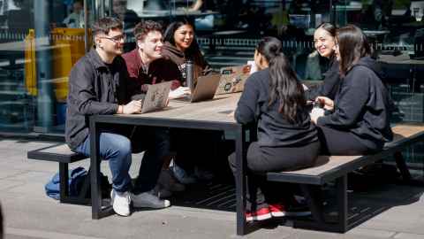 Group of six students sitting at a table outside a building on a sunny day.